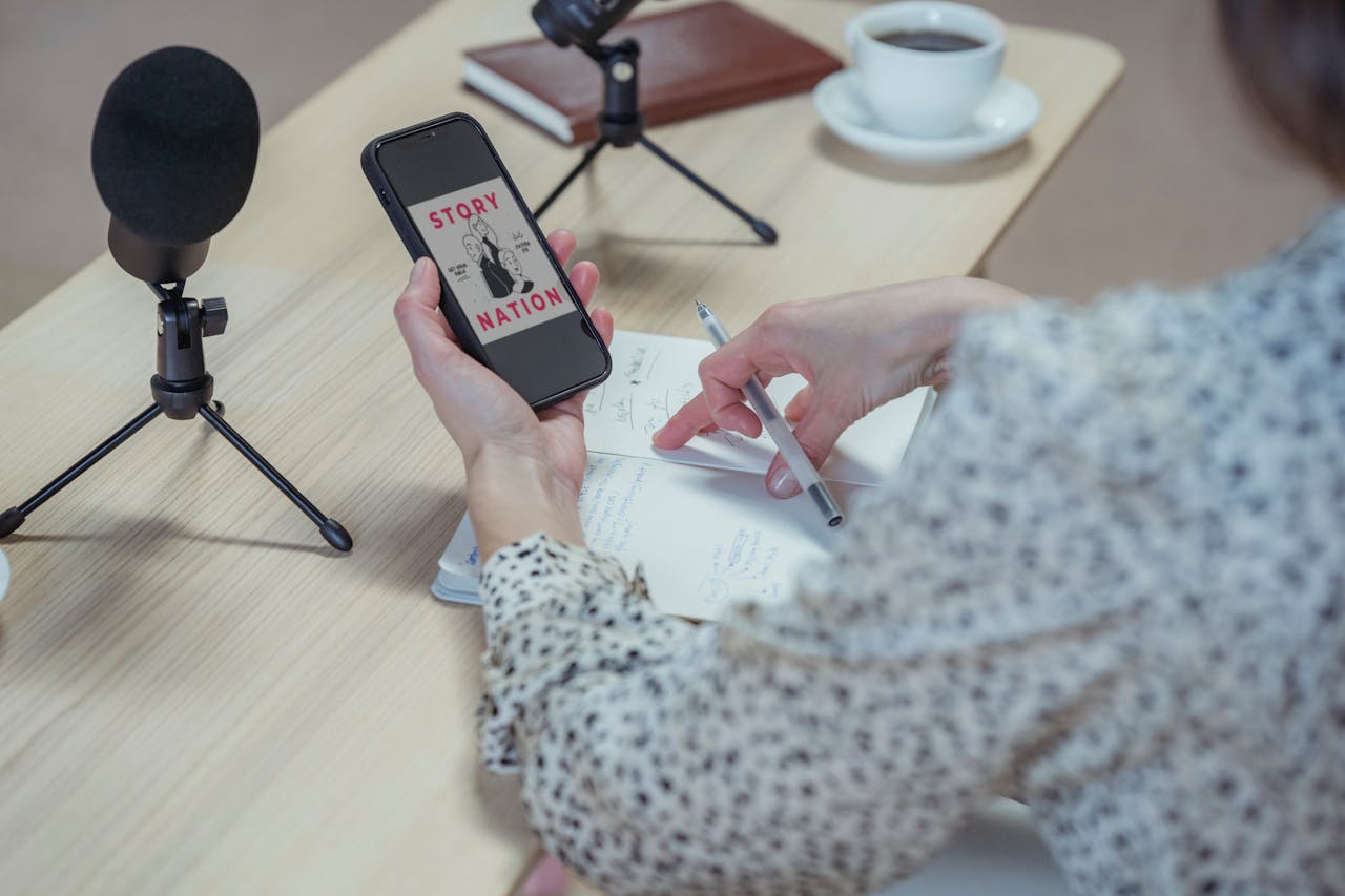 A woman records a podcast at a desk using a smartphone and microphones.